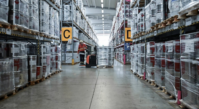 Sayerlack warehouse with stacked metal coating containers and worker using pallet jack in central aisle.