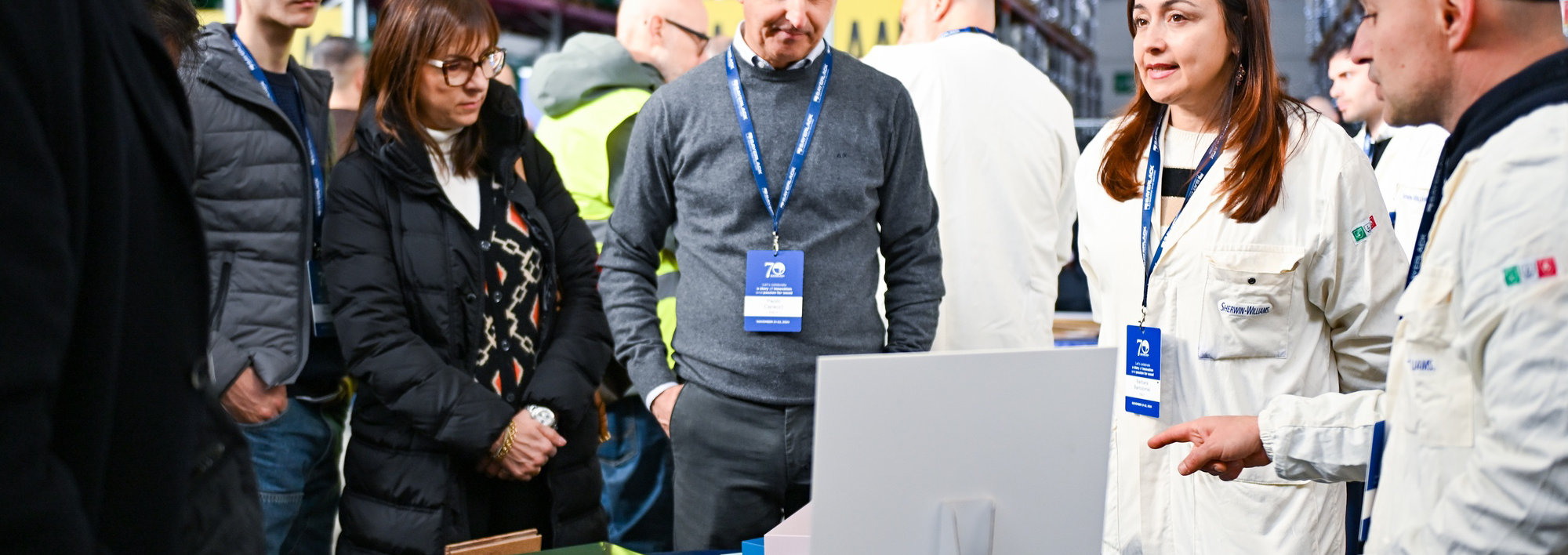 People gathered around a table displaying wood samples and color panels during Sayerlack’s 70th anniversary event in a warehouse setting.