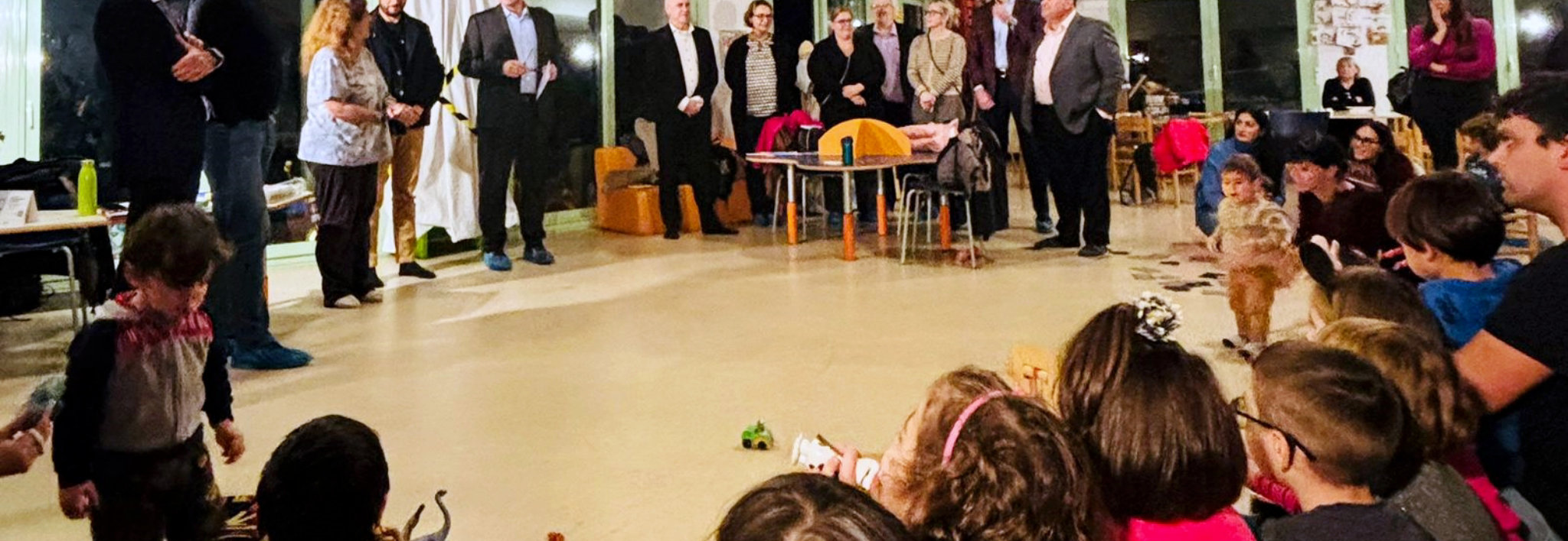 Classroom scene with adults standing and children seated on the floor during an event.