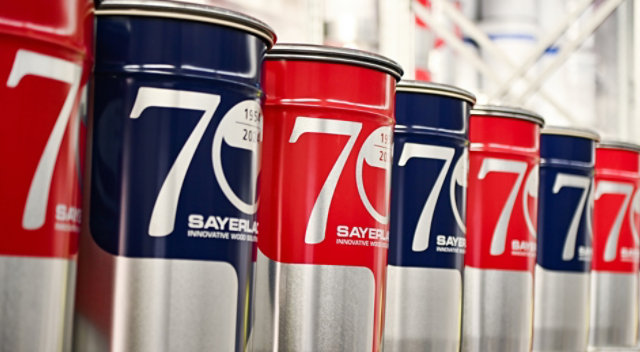 Red and blue Sayerlack paint cans with '70' and '1954–2024' labels, lined up on a shelf.