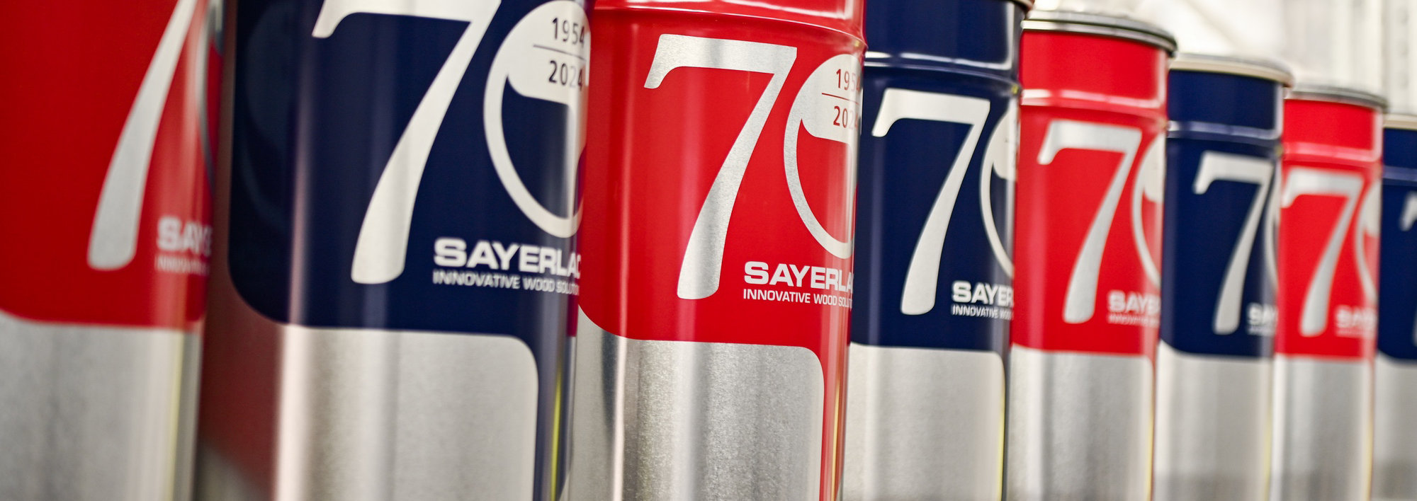 Red and blue Sayerlack paint cans with '70' and '1954–2024' labels, lined up on a shelf.