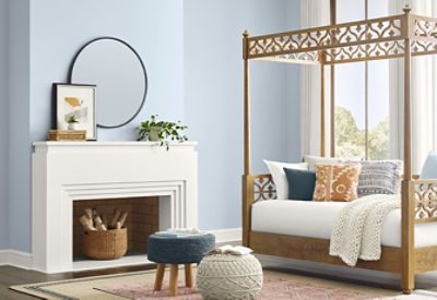 Home interior with light blue-gray walls, painted white fireplace, small fabric stool and pouf in foreground next to wooden canopy daybed piled with accent pillows and draped with knitted white blanket.