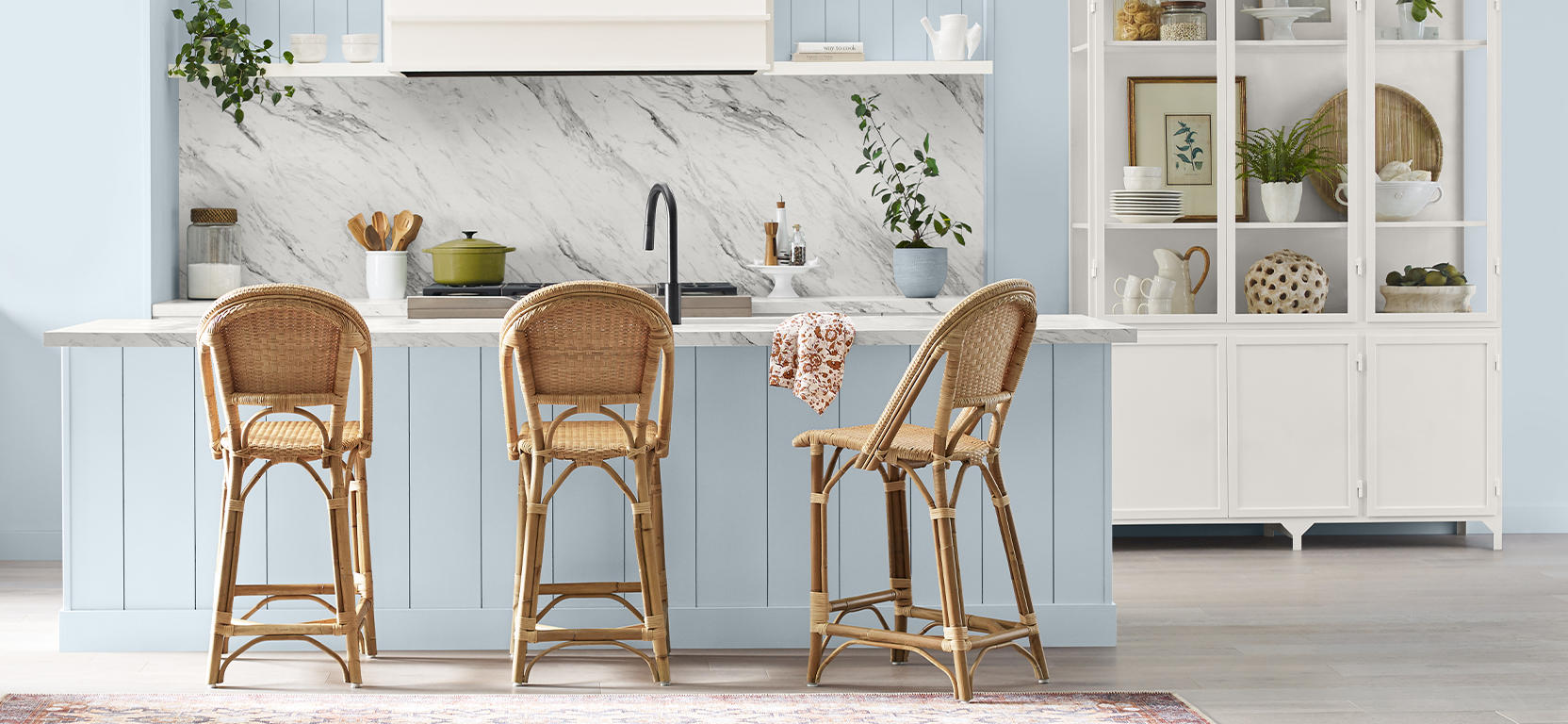 Kitchen with walls and island painted in the Sherwin-Williams Color of the Year, Upward, with rattan chairs, a white marble backsplash, and white cabinet displaying serving pieces and decorative accents.