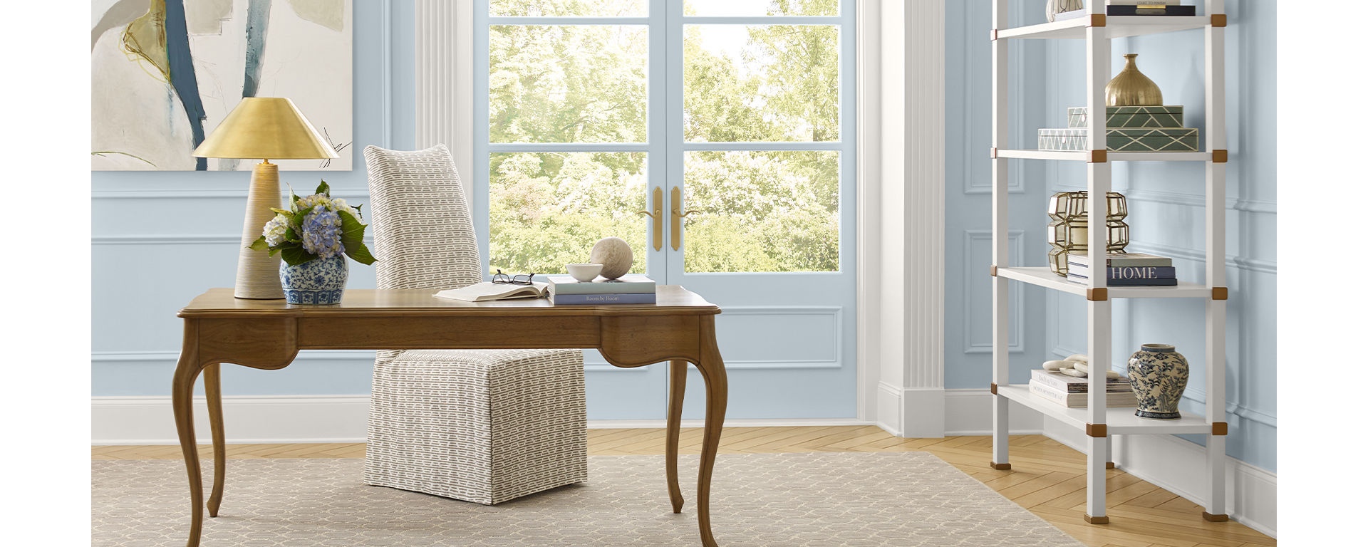 Home office featuring walls painted light blue, fabric-covered chair, wood desk with curved legs, neutral rug on herringbone wood floor, decorative white bookshelf, and French doors to outside.
