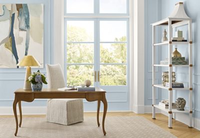 Home office featuring walls painted light blue, fabric-covered chair, wood desk with curved legs, neutral rug on herringbone wood floor, decorative white bookshelf, and French doors to outside.