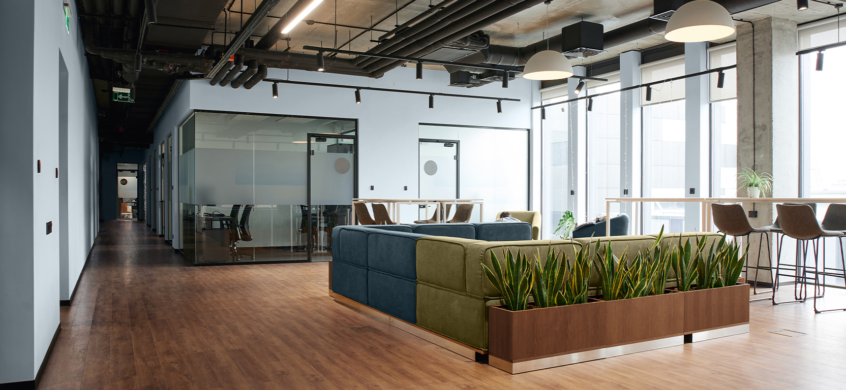 Open concept commercial interior with blue and green sectional seating in center, bar-height tables and chairs surrounding, greenery, high ceilings, industrial styling and wood floors complementing Upward walls.