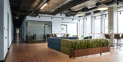 Open concept commercial interior with blue and green sectional seating in center, bar-height tables and chairs surrounding, greenery, high ceilings, industrial styling and wood floors complementing Upward walls.