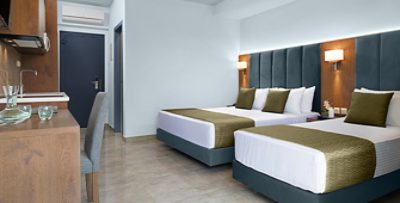 Hotel room with two beds, dark blue velvet headboards and accents with walls painted Upward and medium-stained wood built-in desk and cabinetry.