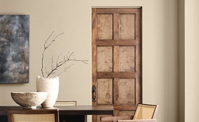 Interior with a dark wooden table, light wooden chairs, ceramic bowl, and vase with branches in front of a Universal Khaki wall and wooden door.