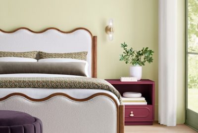 A modern bedroom featuring Celery green walls, a white upholstered scalloped bedframe, a nightstand painted Sherwin-Williams Kirsch Red, and natural light from a large window with trim painted dark Artichoke green.