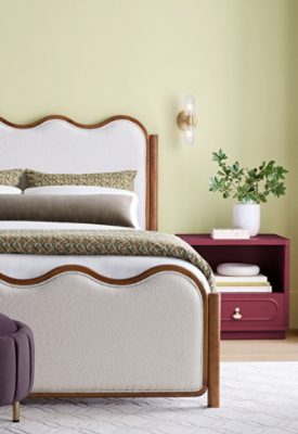 A modern bedroom featuring Celery green walls, a white upholstered scalloped bedframe, a nightstand painted Sherwin-Williams Kirsch Red, and natural light from a large window with trim painted dark Artichoke green.