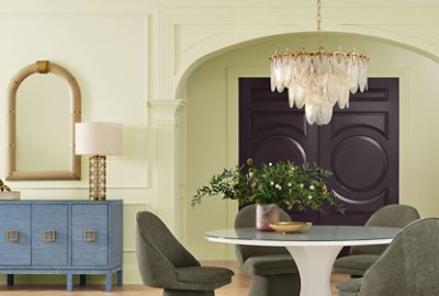 A luxurious dining room featuring Celery walls and Plum Brown double doors, a round white table with upholstered chairs, a blue sideboard with a lamp, and an ornate chandelier overhead.