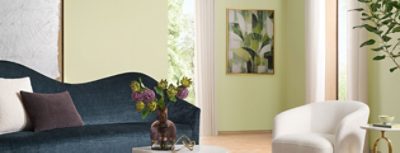 Modern living room with a blue sofa, white chair, purple flowers in a vase, and abstract leaf artwork on a wall painted the Sherwin-Williams Color of the Year, Celery.