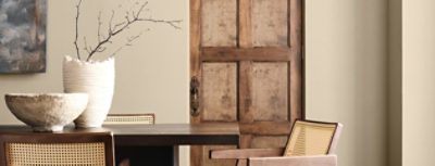 Interior with a dark wooden table, light wooden chairs, ceramic bowl, and vase with branches in front of a Universal Khaki wall and wooden door.