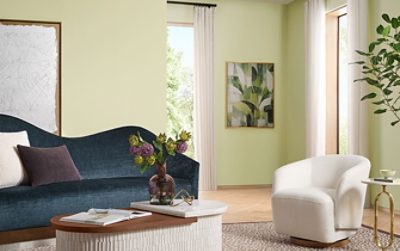 Modern living room with a blue sofa, white chair, purple flowers in a vase, and abstract leaf artwork on a wall painted the Sherwin-Williams Color of the Year, Celery.
