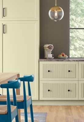 A modern kitchen featuring Celery cabinets, Cocoon walls, a gray marble countertop with an espresso maker and fruit bowl, and Georgian Bay chairs at a wooden dining table with a vase of green plants.