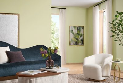 Modern living room with a blue sofa, white chair, purple flowers in a vase, and abstract leaf artwork on a wall painted Sherwin-Williams Color of the Year, Celery.