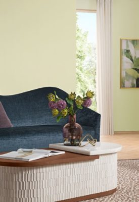 Modern living room with a blue sofa, white chair, purple flowers in a vase, and abstract leaf artwork on a wall painted Sherwin-Williams Color of the Year, Celery.