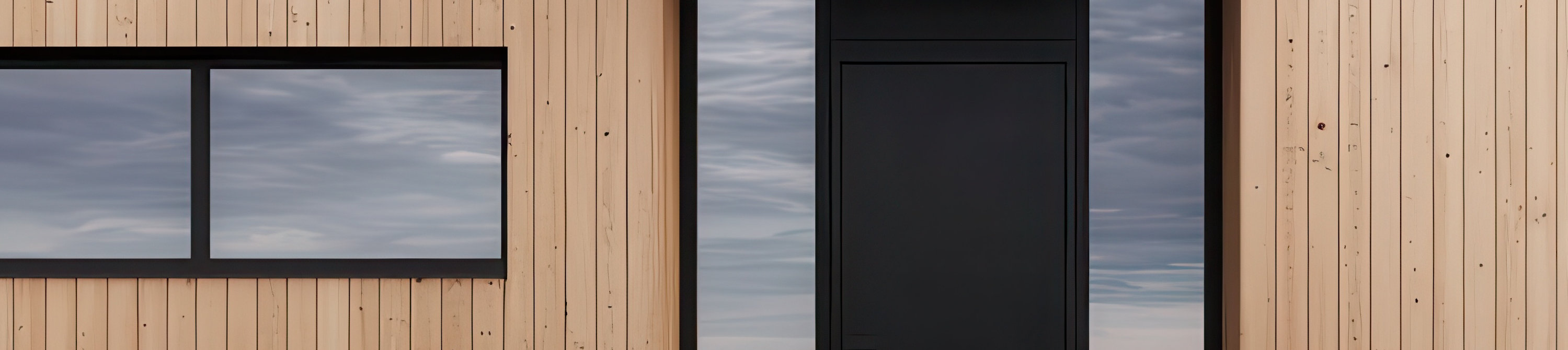 Minimalist wooden design featuring untreated wood walls, blackframed windows, and a gravel path leading to the entrance
