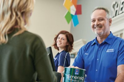 Smiling Sherwin-Williams' store employees passing a can of Emerald paint to a customer.