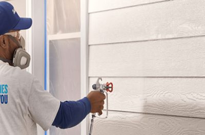 A person wearing safety goggles and a mask applying paint to exterior siding with a spray gun.