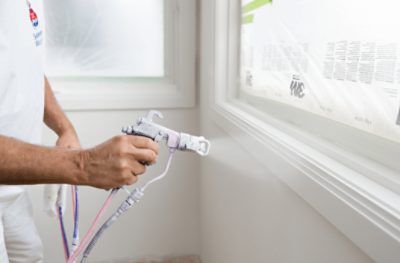 A person using a spray gun to apply paint to interior window trim.