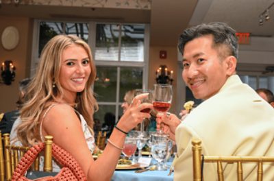 Two well-dressed individuals seated at a table in a social setting, each holding a wine glass and looking directly at the camera.