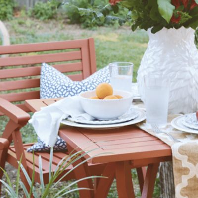 An outdoor table and chair with a vase of flowers and a bowl of lemons on top of two nested plates.