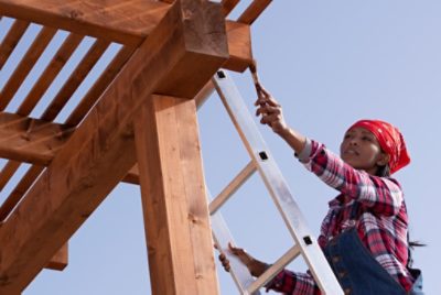 A person on a ladder applying stain to a pergola with a brush.