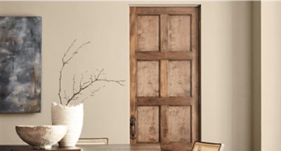 Interior with a dark wooden table, light wooden chairs, ceramic bowl, and vase with branches in front of a Universal Khaki wall and wooden door.