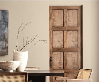 Interior with a dark wooden table, light wooden chairs, ceramic bowl, and vase with branches in front of a Universal Khaki wall and wooden door.