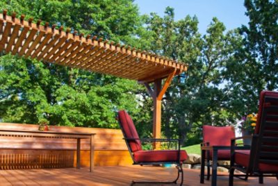 A stained pergola as part of a deck with lush green trees behind.