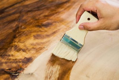 Close-up of a person applying dark wood stain to a wooden surface using a brush.