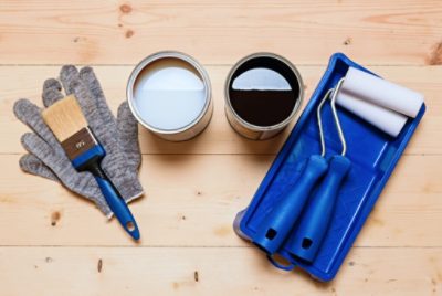 Top-down view of painting tools including a brush resting on top of gloves, rollers in a small paint pan, and cans of white paint and wood stain placed in the center on a light wooden floor.