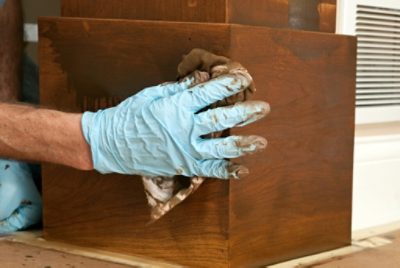 Close-up of a person wearing gloves while applying wood stain with a cloth, highlighting improper application, product waste, and uneven finish.