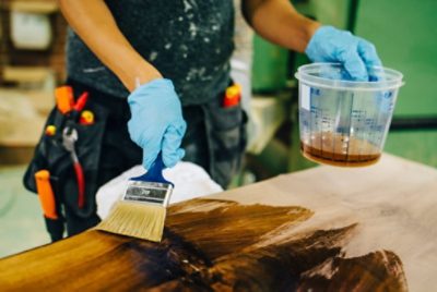 Person wearing gloves holding a stain container and applying heavy coats with a large brush, showing overuse and uneven finish.