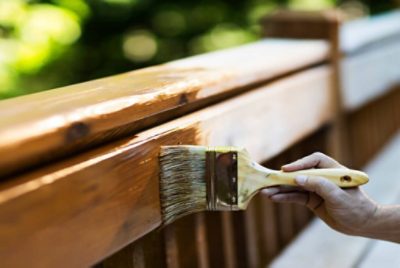 Close-up of a person applying wood stain to a wooden railing using a large brush.