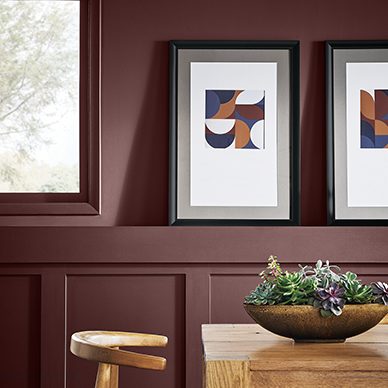 A dark red dining room painted Rookwood Dark Red SW 2801 with a light wood farm table and succulent centerpiece. Two black framed abstract art pieces sit on the chair rail next to the window.