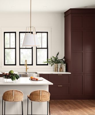 A beige kitchen painted Downing Sand SW 2822 with dark red cabinets, marble countertops, and an island painted a light gray. There are fruits and vegetables on the island and two wicker, low backed chairs next to it.