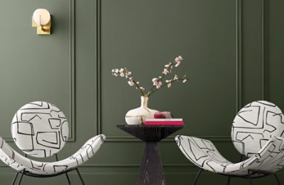 A dark green living room with paneled walls painted dark green Rosemary SW 6186 with two modern white chairs with a black geometric design with a modern accent table between them with books and cherry blossoms in a vase on top.