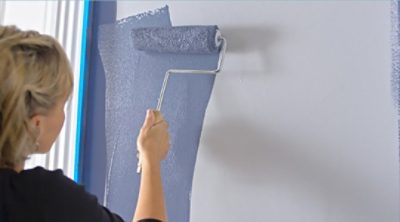 A woman roller painting walls in a room. SW color featured: SW 6243 Distance.