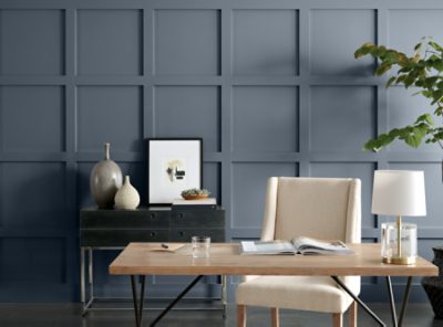 A blue home office with wainscoting walls, light wooden table, and floor plant