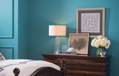 A pale blue bedroom wall with dresser, wall art, and a bed