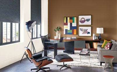 Modern office with one brown wall, one white wall, 2 windows, colorful art pieces, and neutral furnture.