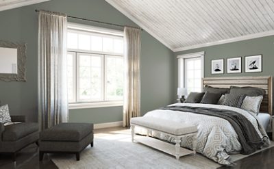 Bedroom with green painted walls, a large window with curtains, chair, bench, and bed made with neutral covers.