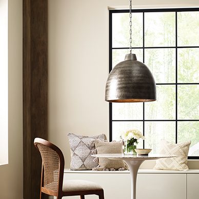 A cozy dining room with a small white metal round pedestal table and one brown chair with a large hammered brass pendant light overhead. The walls are painted cream Slumber Sloth SW 9606 with a white cabinet lining the bottom of the feature wall with a large dark trimmed window.