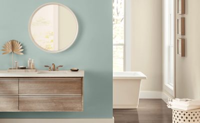 Well lit bathroom with light blue and cream walls, round mirror above a sink countertop and bathtub in the back.