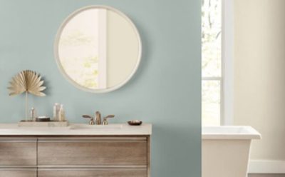 Well lit bathroom with light blue and cream walls, round mirror above a sink countertop and bathtub in the back.