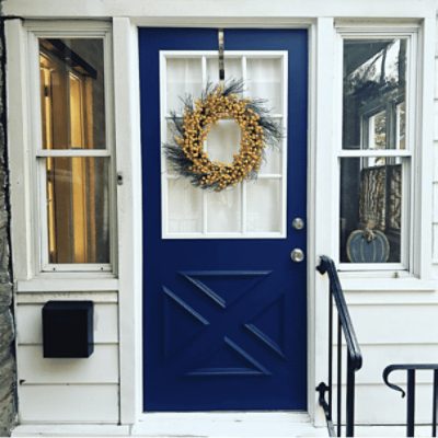 An entrance to a house with white siding, a black mailbox and railing with a blue front door and yellow wreath.