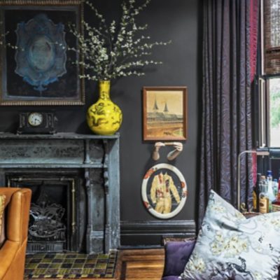 A living room with dark walls and fire place with multi-colored tiles. Various paintings hang from the walls. 